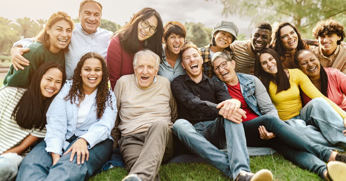 A large, diverse group of multi-generational people sitting close together outdoors in a park, smiling and laughing broadly at the camera