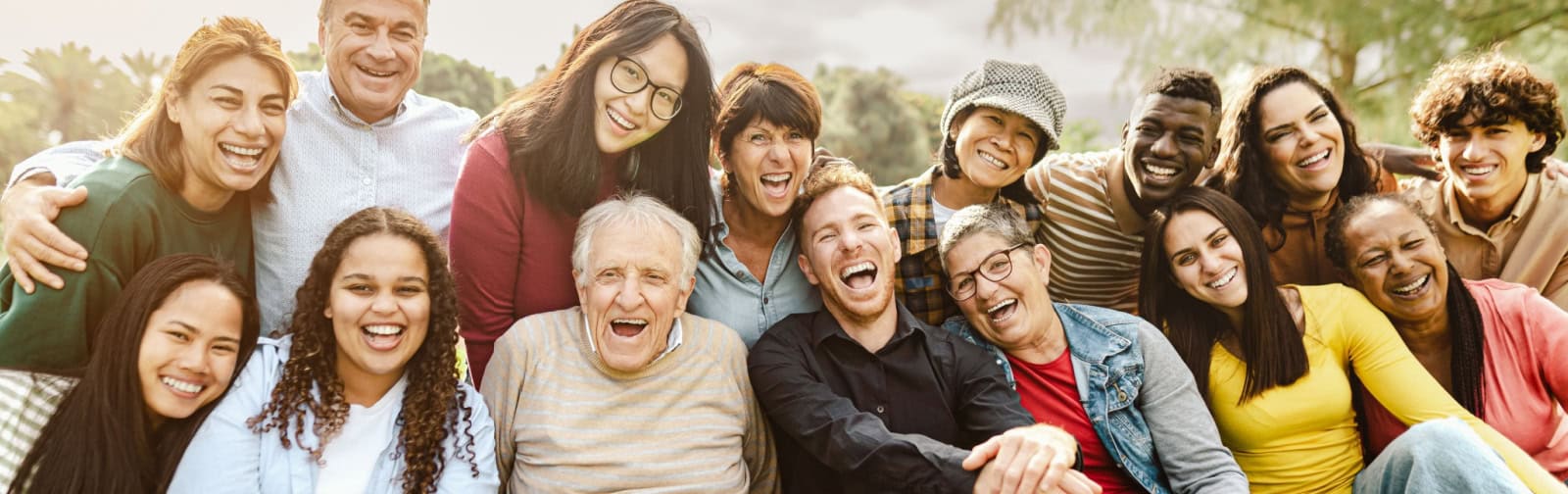 A large, diverse group of multi-generational people sitting close together outdoors in a park, smiling and laughing broadly at the camera